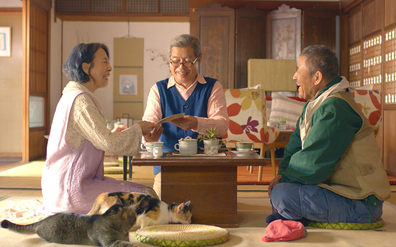 An elderly woman and two men sitting in a Japanese-style room having tea as two cats lounge in the foreground