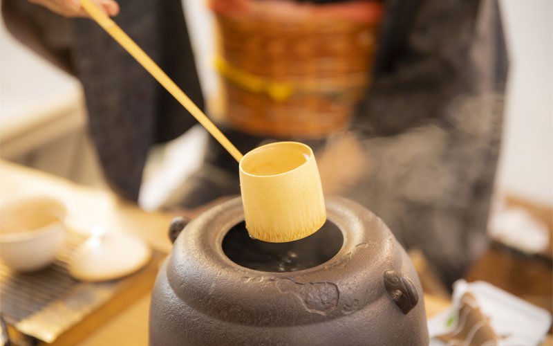 A woman in a kimono scooping hot water out of a pot with a bamboo scoop