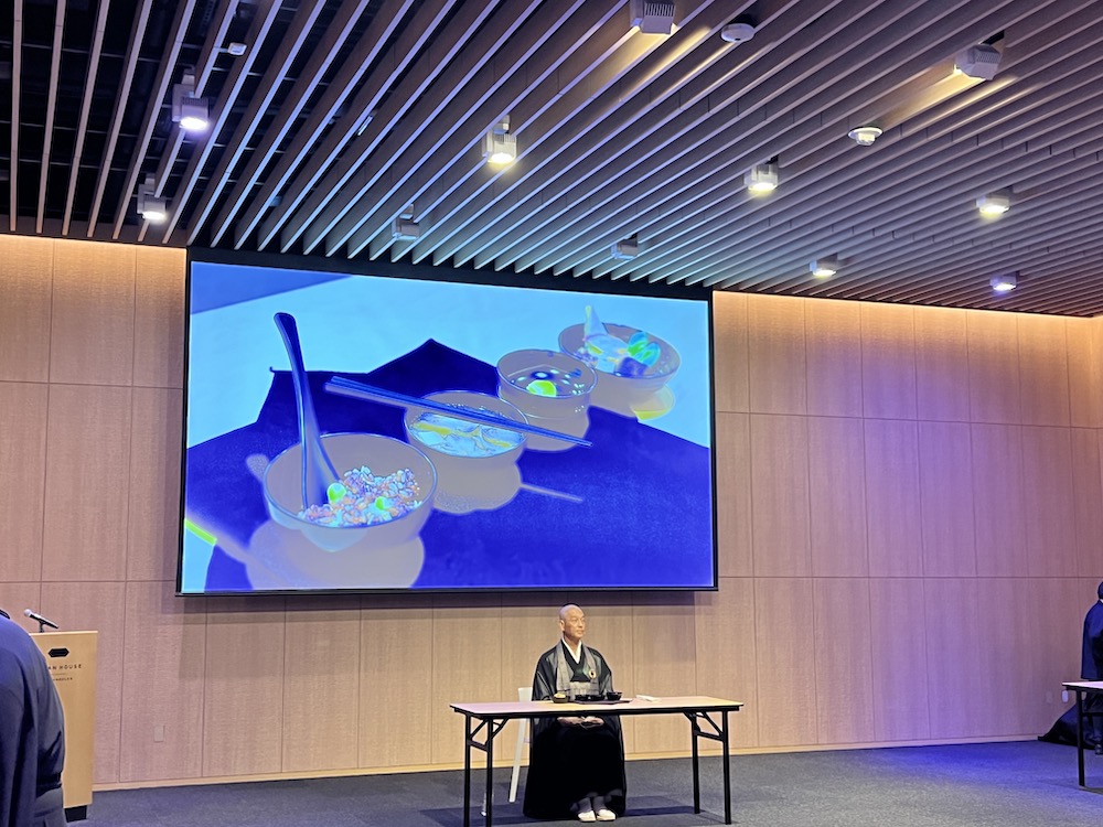 Reverend Kojima sitting at a desk in front of a projection slide about shojin ryori