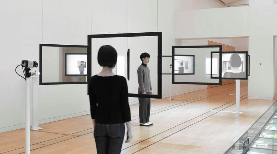 Art installation with mirrors reflecting a person and multiple screens, creating infinite reflections.