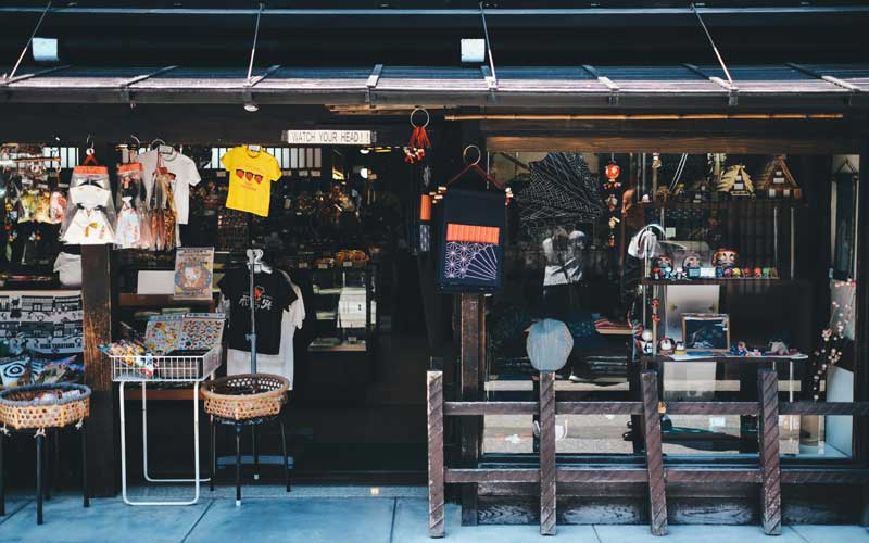 Exterior of a small Japanese souvenir shop with wooden façade, displaying hanging T-shirts, lanterns, textiles, and assorted trinkets at the entrance.