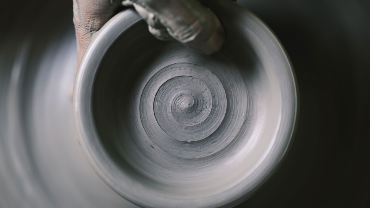 Hands shaping the spiral-textured interior of a gray vessel spinning on a pottery wheel.